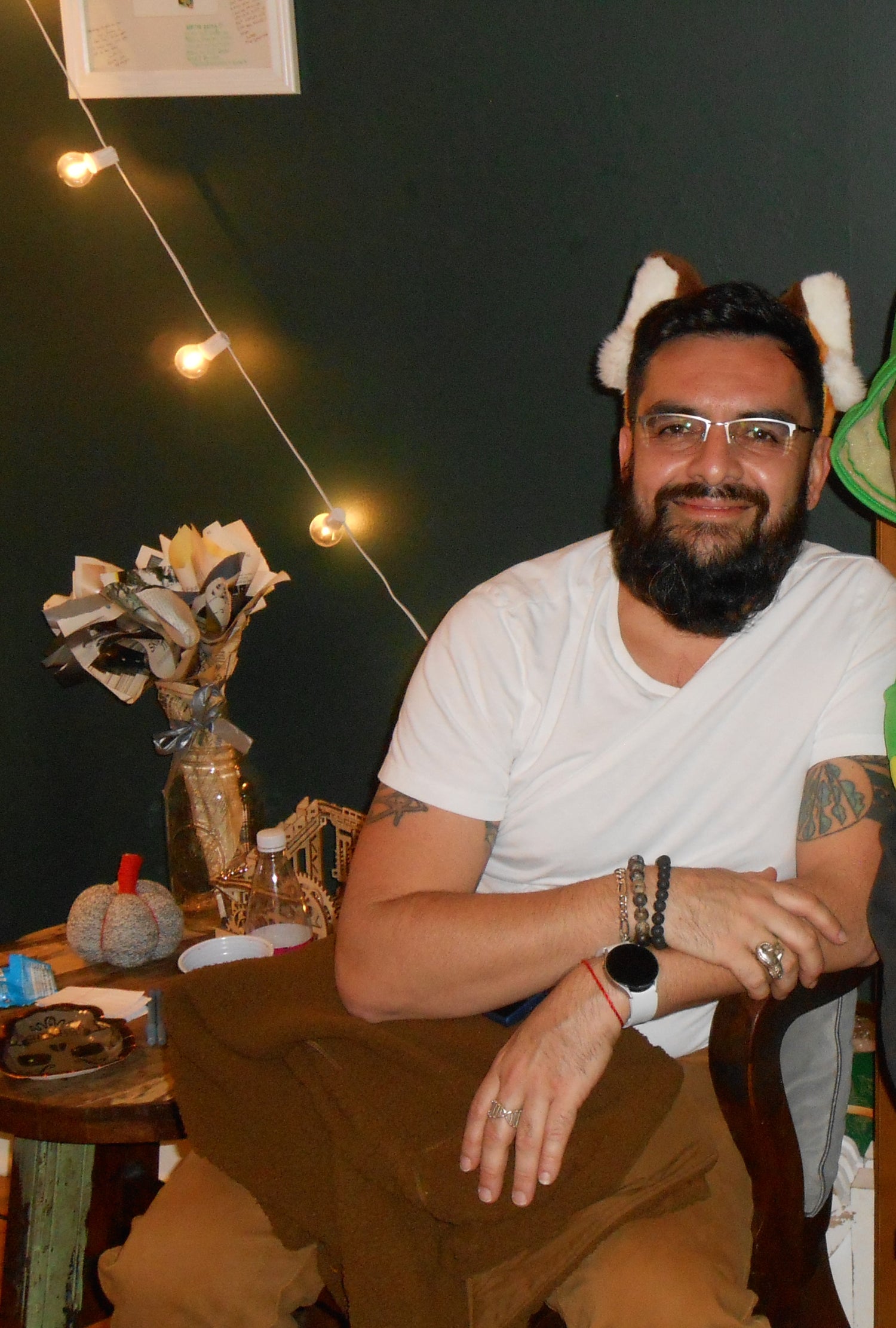 Man with a beard wearing glasses and a white shirt sitting in a casual setting with string lights and decor. He has a pair of red panda ears and glasses.